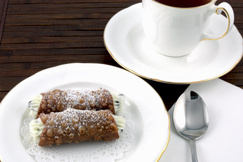 Cannoli Siciliani A close up photo of a teacup and saucer, a silver spoon on a white napkin and cannoli siciliani served on a white plate lined with a white doilie all on a dark wooden placemat.