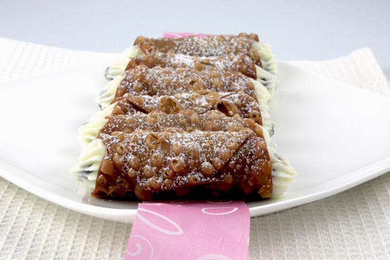 Cannoli Siciliani A photo of Italian, Cannoli Siciliani pastries plated on a white plate with a pink and white ribbon placed on top of a white, waffle patterned tea towel.
