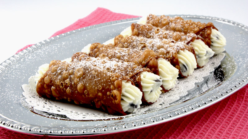 Cannoli Siciliani A close up photo of Cannoli Siciliani (Italian Pastry) on a silver tray lined with white doilies on a pink placemat.