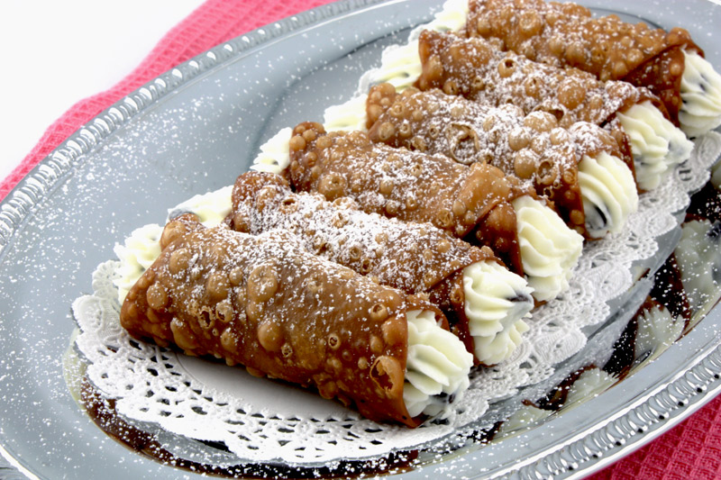 Cannoli Siciliani A close up photo of Cannoli Siciliani (Italian Pastries) on a silver serving tray lined with small white doilies sitting on top of a pink, waffle patterned, tea towel.