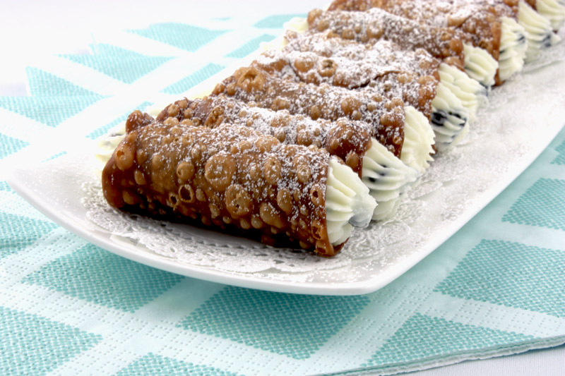 Cannoli Siciliani A close up photo of Cannoli Siciliani (Italian pastries) on a white plate lined with white doilies placed on a bed of blue and white napkins.
