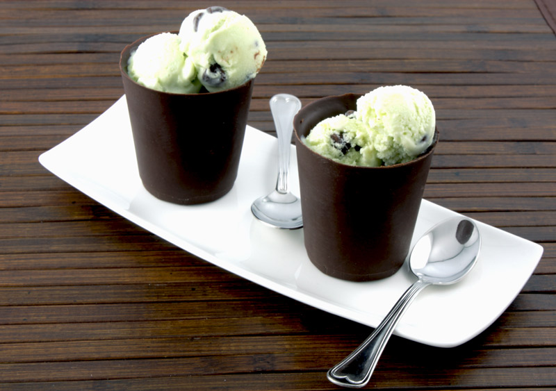 Mint Chocolate Chip Ice Cream A photo of mint chocolate chip ice cream in 2 edible chocolate cups on a white plate with 2 spoons on a wooden placemat.