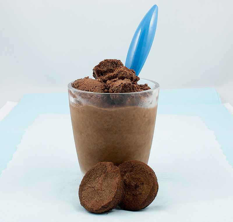 frozen treat recipes, recipe for Donna Hay's double chocolate brownie semifreddo, Italian semifreddo recipes A photo of a cup of double chocolate brownie semifreddo garnished with chopped brownies, with a blue spoon inserted in cup and 2 small brownie bites displayed in front of glass on a piece of white parchment that is sitting atop a blue napkin.