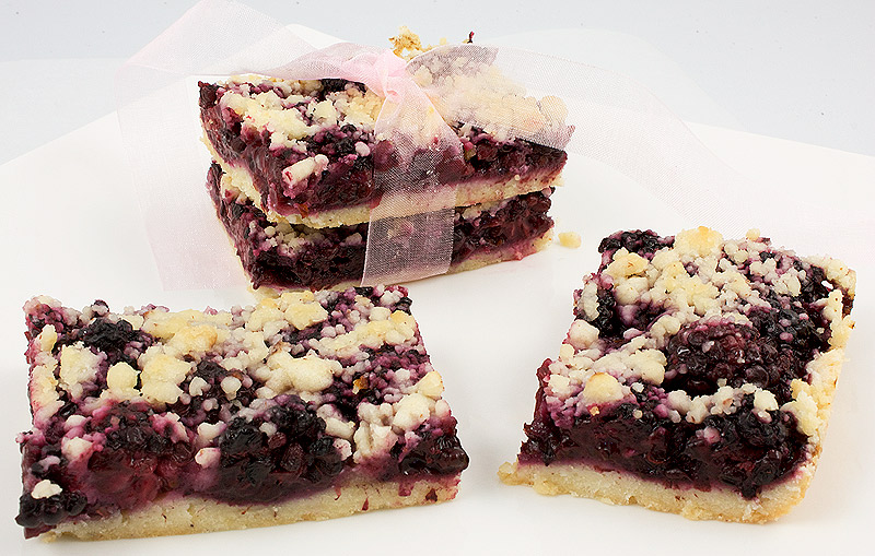 easy recipe for berry crumb bars, easy recipe for blackberry-almond shortbread squares, easy summer desserts, easy summer berry desserts A photo of 4 blackberry-almond shortbread squares, 2 are stacked and tied with a sheer ribbon, all placed on a white dish.