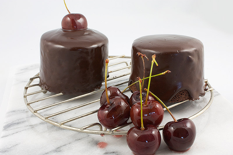 how do I tell when my cake is ready, Donna Hay's tips for baking the perfect cake, chocolate mud cake recipe A photo of 2 individual sized chocolate mud cakes with chocolate ganache glaze with sweet glazed cherries on a wire rack.