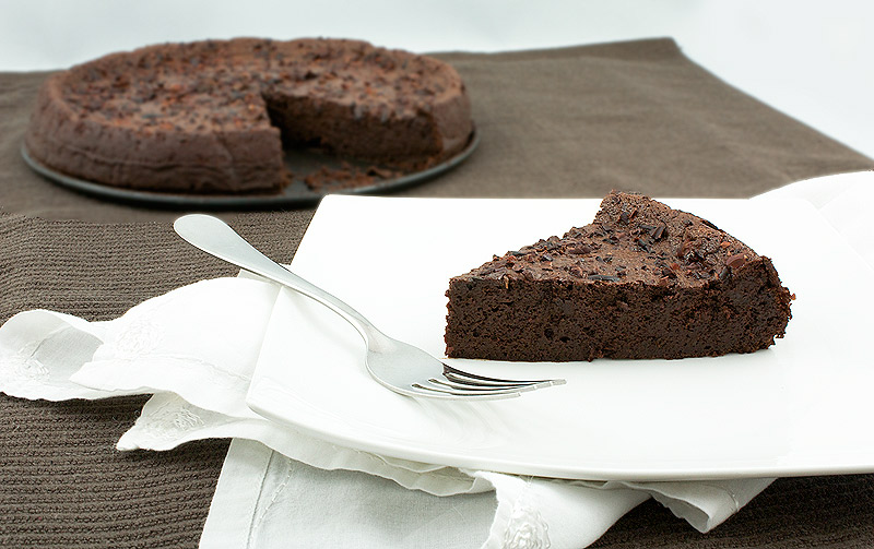 french dessert recipes, flourless chocolate torte, recipe for flourless chocolate cake A photo of a chocolate flourless cake and a square, white plate with a slice of flourless chocolate cake with a dessert fork placed on a white cloth napkin.
