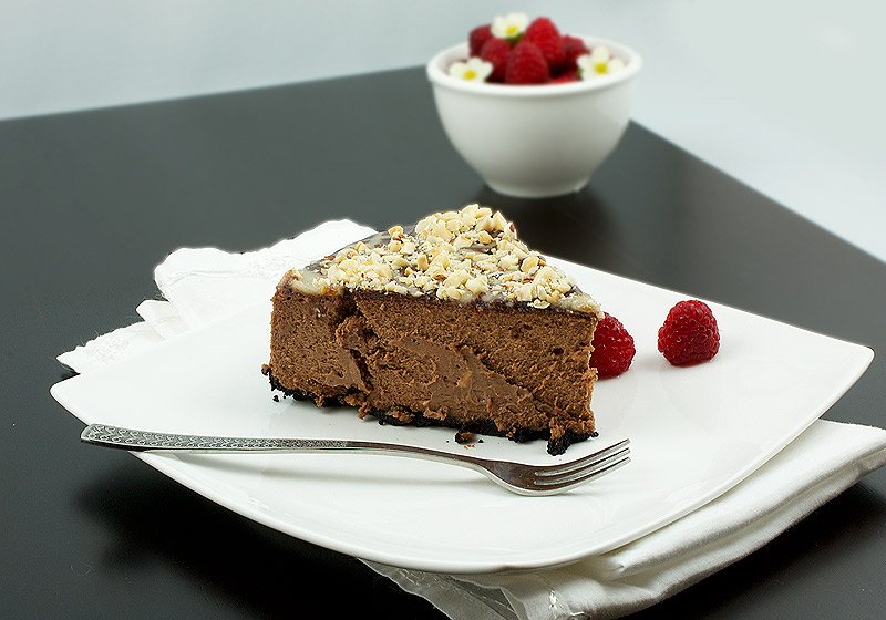 chocolate dessert, dessert recipes with pictures, baked cheesecake recipe A photo of a slice of chocolate cheesecake with Oreo cookie crust topped with chocolate sauce, caramel sauce and toasted hazelnuts on a plate served with a few fresh raspberries.