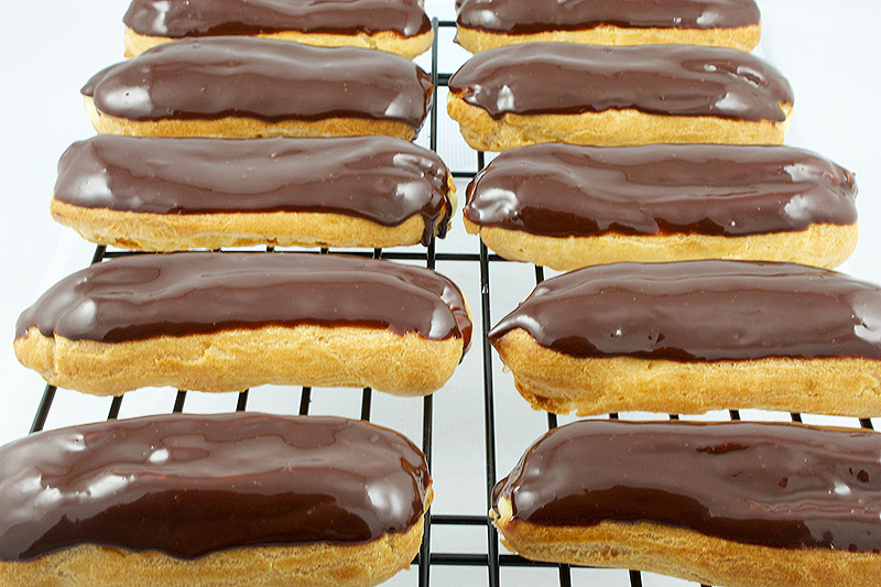 Chocolate Éclairs with Vanilla Bean Crème Pâtissière and Chocolate Ganache Glaze A photo taken looking down on Chocolate Éclairs with Vanilla Bean Crème Pâtissière and Chocolate Ganache Glaze french pastries on a wire rack.