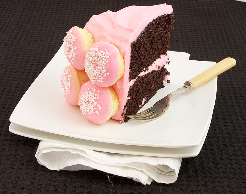 chocolate layer cake recipe, chocolate layered cake, double layer chocolate cake A photo of a slice of Chocolate Layer Cake with White Chocolate Cream Cheese Frosting decorated with mini Baked White Chocolate Doughnuts with pink Vanilla Glaze and sprinkled with white nonpareils on a white dish with a dessert fork.
