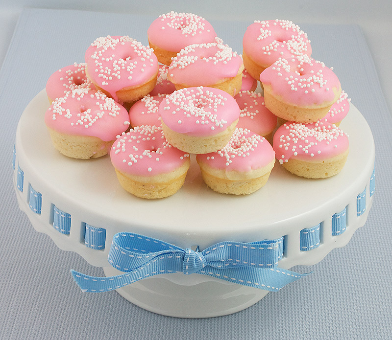 donut recipes, baked doughnuts, doughnut recipes A photo of Baked White Chocolate Cake Doughnuts with pink Vanilla Doughnut Glaze decorated with white nonpareils on a white cake stand.