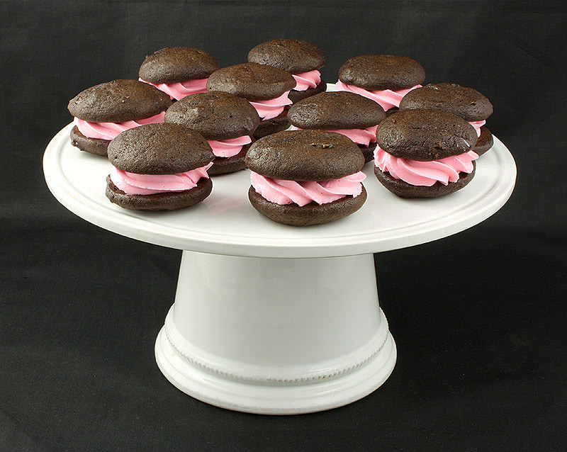 Chocolate Whoopie Pies with Vanilla Swiss Meringue Buttercream A photo of Chocolate Whoopie Pies with Vanilla Swiss Meringue Buttercream Cookies arranged in a single layer on a white cake stand.