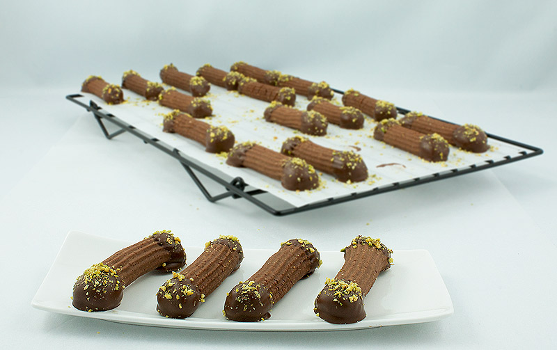 easy Christmas cookies, buttery spritz cookies, spritz cookie recipe A photo of Spritz Cookies dipped in chocolate and sprinkled with pistachio crumb a small plate and in the background a cooling rack with Chocolate Hazelnut Spritz Cookies.
