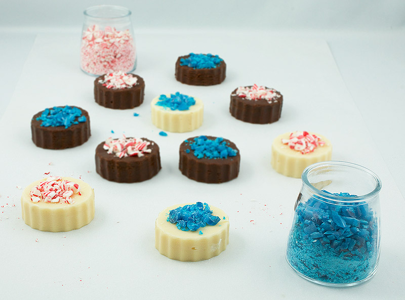 fudge candy recipes, white chocolate recipes, chocolate fudge recipes A photo of various pieces of Holiday Fudge decorated with crushed candy canes randomly placed on a piece of parchment and 2 containers of crushed candy canes.