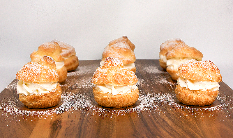 Profiteroles with Vanilla Crème Chantilly A photo of Profiteroles with Vanilla Creme Chantilly dusted with confectioners' sugar on a wooden cutting board.