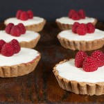 Hazelnut, Raspberry Jam and White Chocolate Ganache Tarts A photo of six Hazelnut, Raspberry Jam and White Chocolate Ganache Tarts on a dark wooden board.