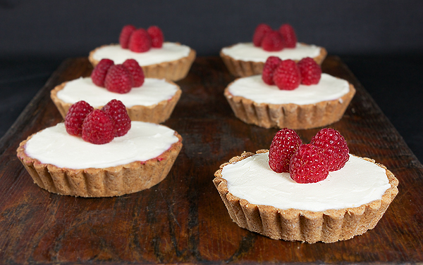 Hazelnut, Raspberry Jam and White Chocolate Ganache Tarts A photo of six Hazelnut, Raspberry Jam and White Chocolate Ganache Tarts on a dark wooden board.