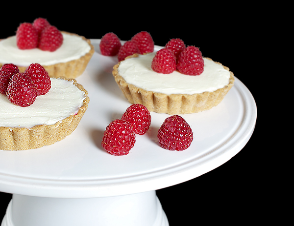 White Chocolate Tarts with Raspberries A photo of White Chocolate Tarts with a layer of seedless Raspberry Jam and garnished with fresh raspberries displayed on a white cake stand.