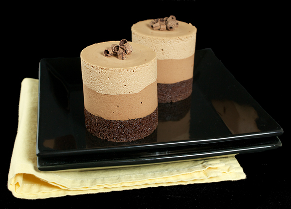 recipe for chocolate mousse cakes, mini cake recipes, best dessert recipes A photo of 2 mini cakes layered with Dark Chocolate Cake, Bittersweet Chocolate Mousse and Milk Chocolate Mousse on a black dessert dish with a black background.
