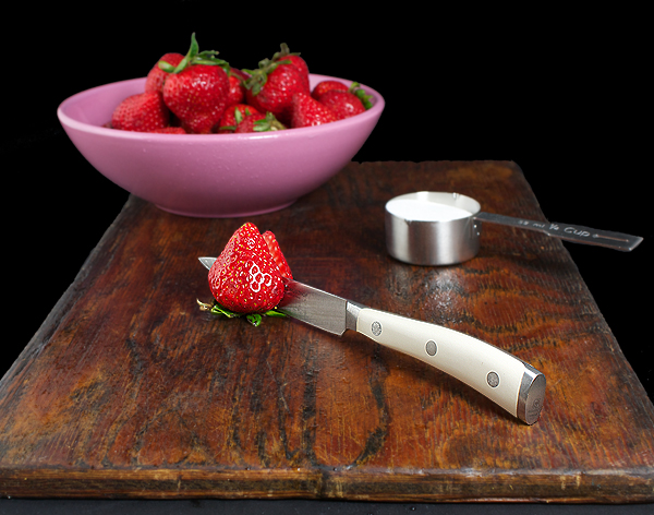 Strawberry Shortcake Recipe A photo of a bowl of fresh strawberries and sugar displayed on a dark wooden board.
