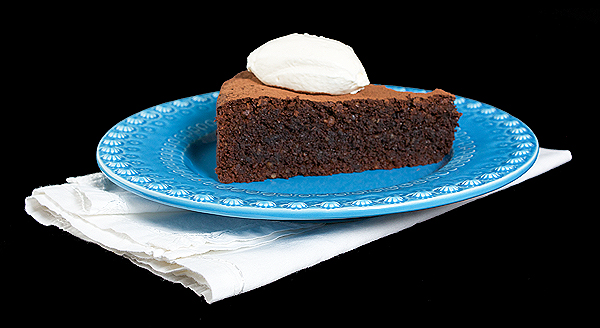 flourless chocolate cakes, recipe for torte, how to make a chocolate cake A photo of a slice of Torta Caprese with a quenelle of Sweetened Whipped Cream and a dusting of cocoa placed on a blue dessert dish.