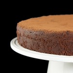 Torta Caprese - Flourless Chocolate Torte A cropped photo of an Italian Torta Caprese dusted with cocoa and displayed on a white cake stand.