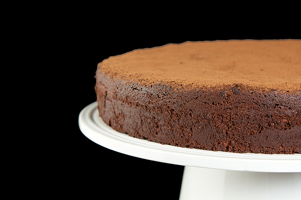 Torta Caprese - Flourless Chocolate Torte A cropped photo of an Italian Torta Caprese dusted with cocoa and displayed on a white cake stand.
