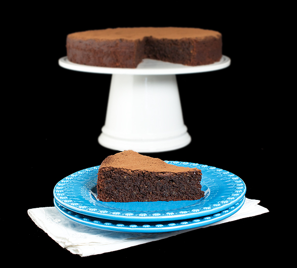 Italian Cake A photo of Italian Cake, Torta Caprese displayed on a white cake stand with a slice removed and displayed on a blue dessert dish set in front.