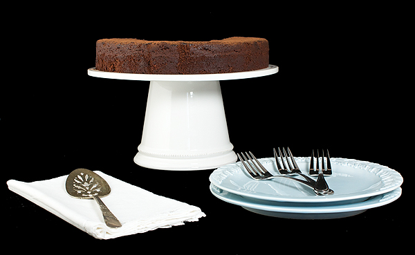 italian cake recipes, italian chocolate, easy chocolate cake A photo of a Torta Caprese displayed on a white cake stand along side pale blue dessert dishes, dessert forks and a vintage cake lifter on a white cloth napkin.