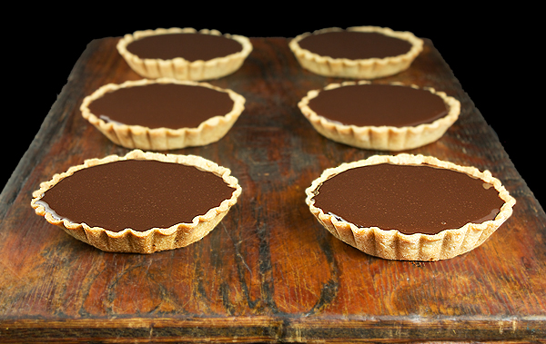 Chocolate Tart A photo of 6 Chocolate Tarts with a layer of caramel and toasted hazelnuts displayed on a dark wooden board.