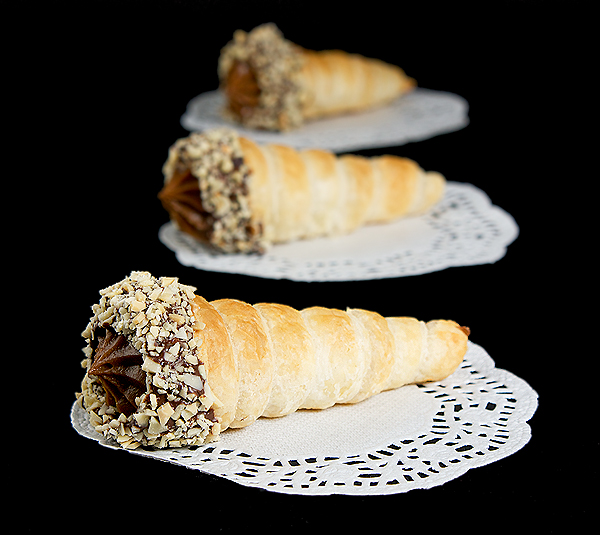 Italian Pastry, Cannoncini di Sfoglia A photo of three Cannoncini di Sfoglia con Crema Pasticcera al Cioccolato (Pastry Horns filled with Chocolate Pastry Cream) place on white doilies and displayed in a row, one behind the other.