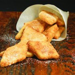 Grace's Sweet Life Crema Fritta alla Veneziana A photo of Crema Fritta alla Veneziana, Venetian Style Fried Cream, dusted with confectioners' sugar in a paper cone displayed on a dark wooden board.