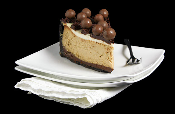 Grace's Sweet Life Mocha Cheesecake A photo of a slice of Mocha Cheesecake decorated with Kahlúa-Chocolate Ganache in a lattice design and garnished with chocolate whoppers on a white dessert dish with a dessert fork.