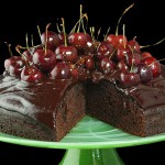 Chocolate Cake with Chocolate Ganache A photo of Chocolate Cake frosted with Chocolate Ganache and decorated with Glazed Cherries with a slice removed and displayed on a pale green frosted glass cake stand.