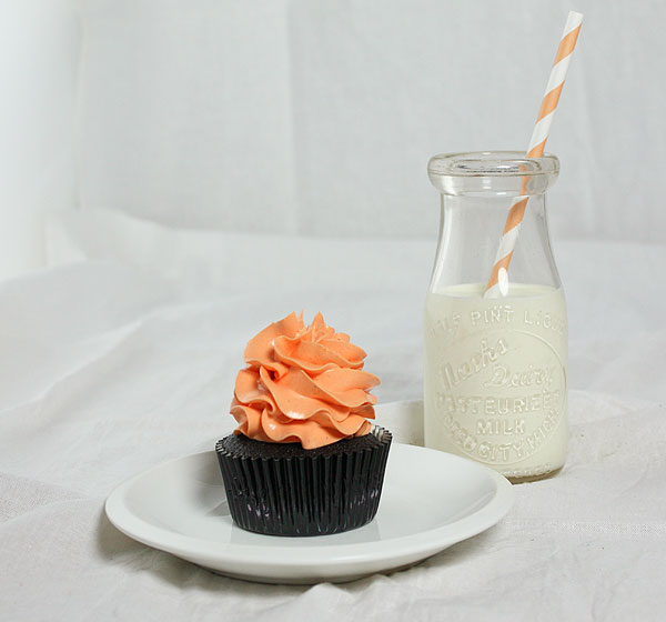 Dark Chocolate Pumpkin Cupcakes from Grace's Sweet Life A photo of a Dark Chocolate Pumpkin Cupcake decorated with orange-tinted Swiss Meringue Buttercream with a small jug of milk with an orange and white striped straw.