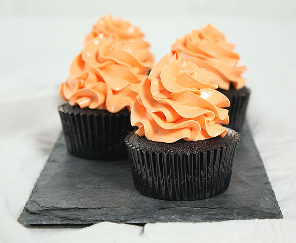Grace's Sweet Life Chocolate Cupcakes with Pumpkin A photo taken of four Chocolate Cupcakes with Pumpkin decorated with orange-tinted frosting displayed in a row on a black slate board.