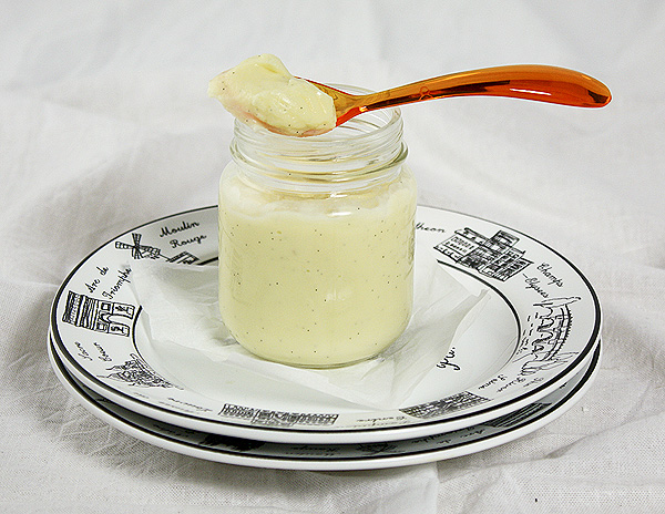 Grace's Sweet Life White Chocolate Pudding Close up photo of White Chocolate Pudding in a glass jar displayed on a stack of dessert dishes with an orange acrylic spoon.