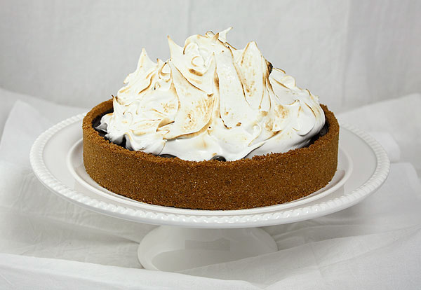 A photo of Baileys S mores Pie displayed on white cake stand with bottle of Baileys and graham crackers.