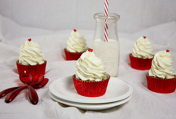 A photo of four Gourmet Strawberry Cupcakes with Swiss Meringue Buttercream each decorated with one red heart sprinkle on top in red foil cupcake liners displayed with milk in a glass bottle with red and white striped straw.