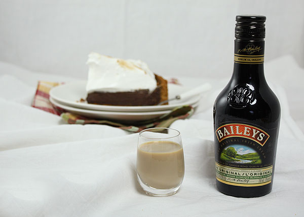 A photo of a slice of Smores Pie on double stack of white dessert dishes displayed with bottle and shot of Baileys.