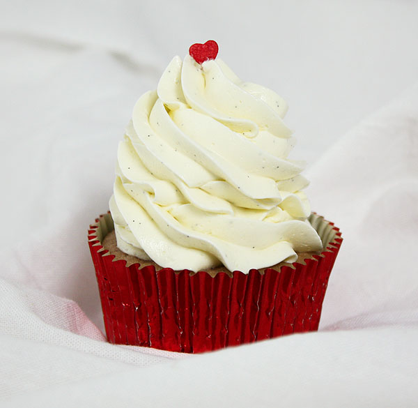 Close up photo of a Strawberry Cupcake with Swiss Meringue Buttercream decorated with a single red heart sprinkle on top in red foil cupcake liner.
