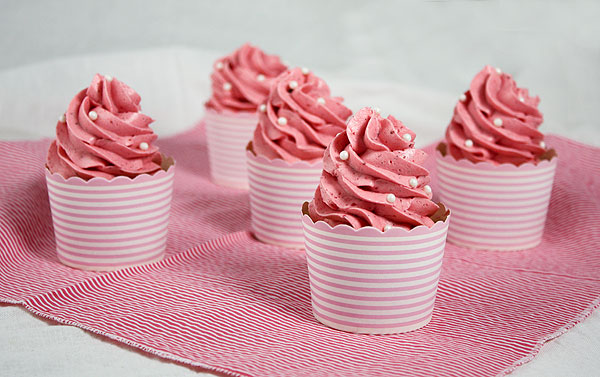 A photo of Strawberry Cupcakes with Pink Strawberry Meringue Buttercream frosting decorated with white sugar pearls displayed on pink and white striped cloth napkin.