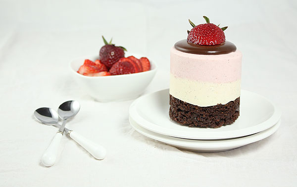 A photo of a Mini Neapolitan Mousse Cake displayed with bowl of fresh strawberries and dessert spoons.