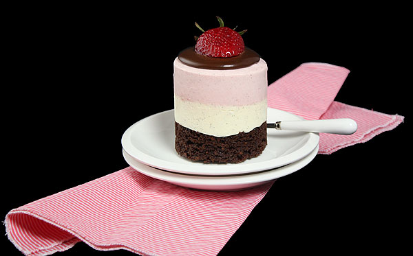A photo of a Mini Neapolitan Mousse Cake plated on stack of two white dessert plates displayed on pink and white striped napkin.