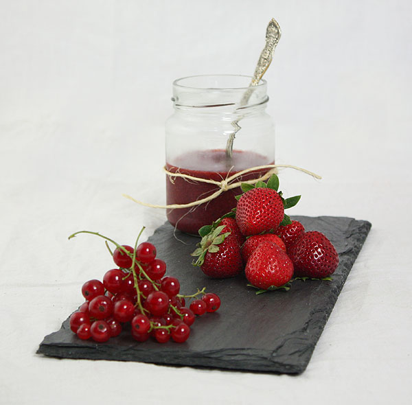 Photo of a jar of Baked Strawberry Jam displayed on slate board with fresh strawberries and red currants.