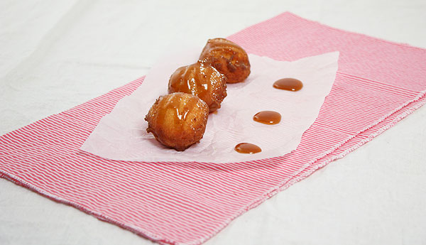 Photo of three Ricotta Beignets drizzled with Butter-Rum Caramel Sauce displayed in a row, one behind the other, on pink and white striped napkin lined with parchment square.