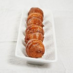 Photo of Sweet Ricotta Fritters dusted with confectioners' sugar and drizzled with butter-rum caramel sauce displayed on white rectangle-shaped serving dish displayed in a row, one behind the other.