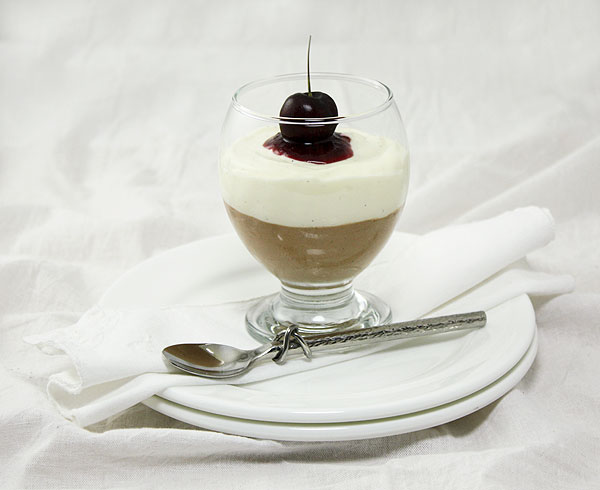 Triple Chocolate Mousse dessert in glass topped with cherry sauce and fresh cherry plated on double stack of white dessert dishes with dessert spoon.
