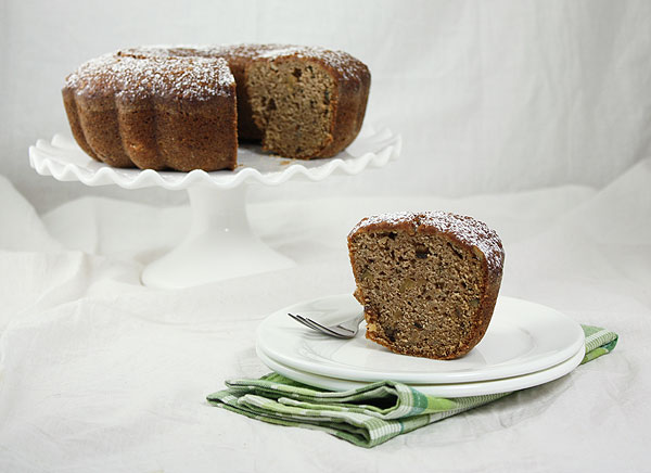 A photo of a slice on Pear Bread displayed on white dessert dish set in front of whole cake displayed on white cake stand.